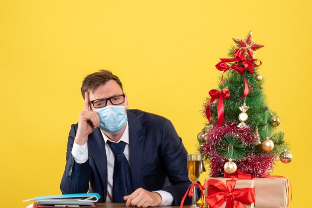 Foto gratuita vista frontal del hombre de negocios deprimido sentado en la mesa cerca del árbol de navidad y presenta en amarillo