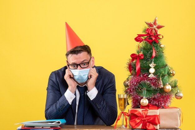 Vista frontal del hombre de negocios deprimido sentado en la mesa cerca del árbol de Navidad y presenta en amarillo