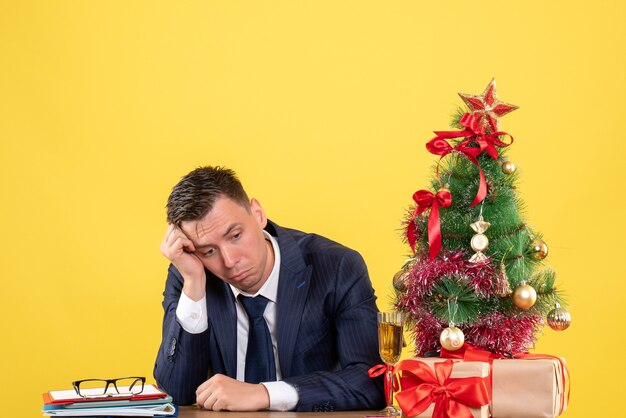 Vista frontal del hombre de negocios deprimido sentado en la mesa cerca del árbol de Navidad y presenta en amarillo