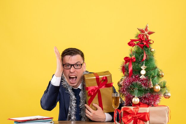 Vista frontal del hombre de negocios enojado sentado en la mesa cerca del árbol de Navidad y presenta en la pared amarilla