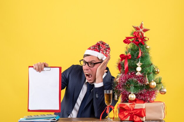 Vista frontal del hombre de negocios enojado sentado en la mesa cerca del árbol de Navidad y presenta sobre fondo amarillo