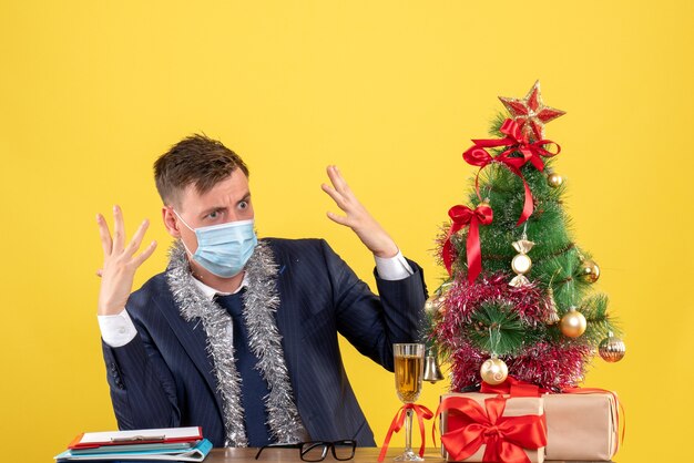 Vista frontal del hombre de negocios enojado sentado en la mesa cerca del árbol de Navidad y presenta sobre fondo amarillo