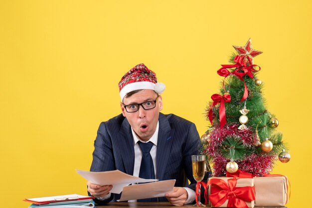 Vista frontal del hombre de negocios feliz revisando papeles sentado en la mesa cerca del árbol de Navidad y presenta en amarillo