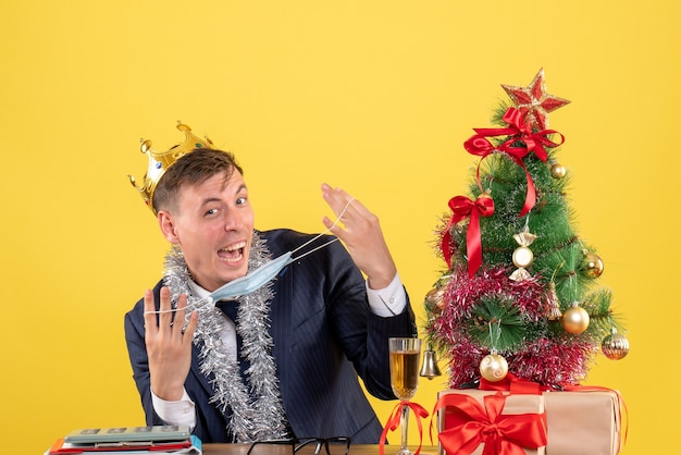 Vista frontal del hombre de negocios feliz sosteniendo su máscara sentado en la mesa cerca del árbol de Navidad y presenta en amarillo