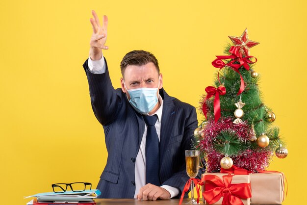 Vista frontal del hombre de negocios gritando a alguien sentado en la mesa cerca del árbol de Navidad y presenta en amarillo