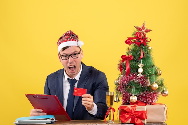 Vista frontal del hombre de negocios gritó sosteniendo el portapapeles sentado en la mesa cerca del árbol de Navidad y presenta en amarillo.