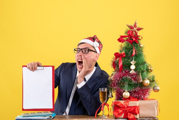 Vista frontal del hombre de negocios gritó sosteniendo el portapapeles sentado en la mesa cerca del árbol de Navidad y presenta en amarillo.