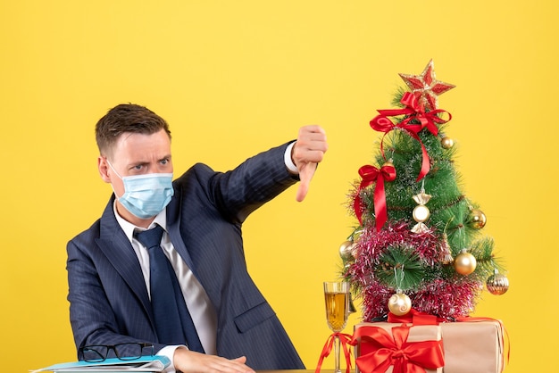 Vista frontal del hombre de negocios haciendo el pulgar hacia abajo firmar sentado en la mesa cerca del árbol de Navidad y presenta en amarillo