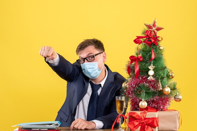 Vista frontal del hombre de negocios levantando la mano sentado en la mesa cerca del árbol de Navidad y presenta en amarillo