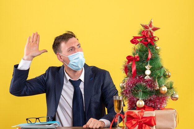 Vista frontal del hombre de negocios levantando la mano sentado en la mesa cerca del árbol de Navidad y presenta en amarillo