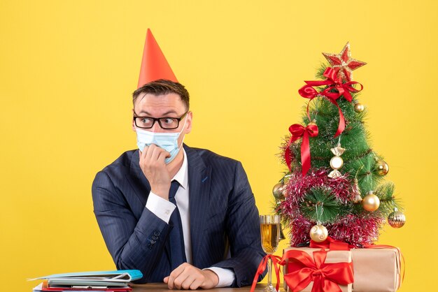 Vista frontal del hombre de negocios con máscara y gorro de fiesta sentado en la mesa cerca del árbol de Navidad y regalos en amarillo