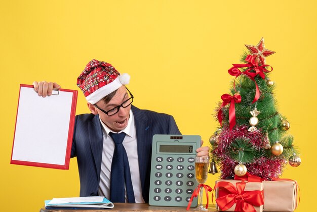 Vista frontal del hombre de negocios mirando la calculadora sentado en la mesa cerca del árbol de Navidad y presenta en amarillo