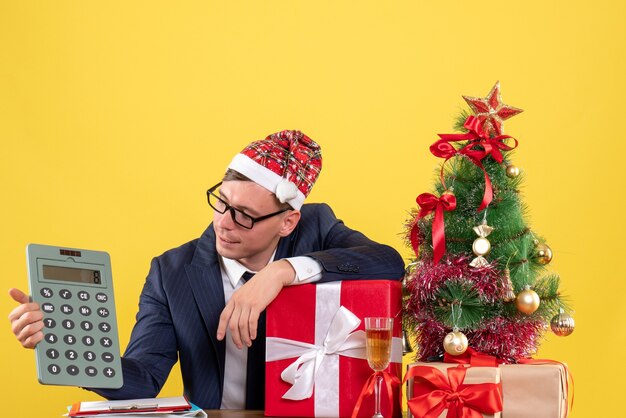 Vista frontal del hombre de negocios mirando su calculadora sentado en la mesa cerca del árbol de Navidad y presenta en amarillo