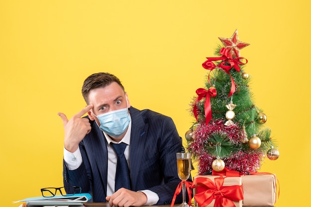 Vista frontal del hombre de negocios poniendo la pistola en la sien sentado en la mesa cerca del árbol de Navidad y presenta en amarillo