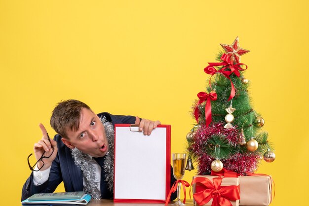 Vista frontal del hombre de negocios con portapapeles y anteojos sentado en la mesa cerca del árbol de Navidad y presenta en amarillo