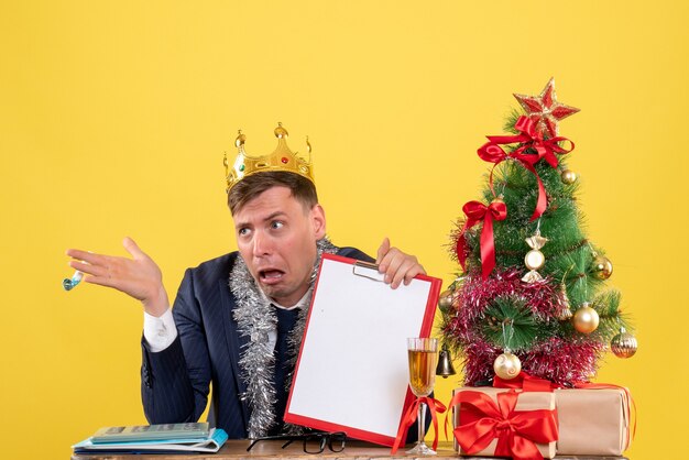 Vista frontal del hombre de negocios con portapapeles y matraca sentado en la mesa cerca del árbol de Navidad y presenta en amarillo