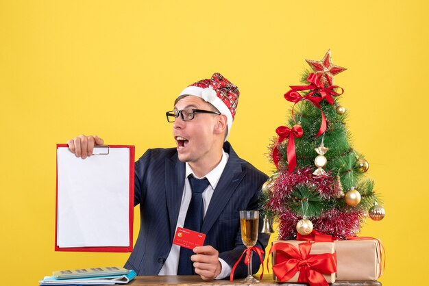 Vista frontal del hombre de negocios con portapapeles y tarjeta sentado en la mesa cerca del árbol de Navidad y regalos en amarillo