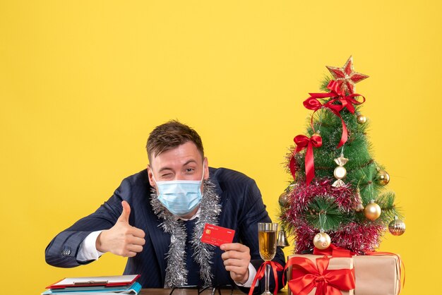 Vista frontal del hombre de negocios que hace el pulgar hacia arriba firmar sentado en la mesa cerca del árbol de Navidad y presenta en amarillo