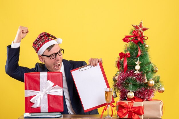 Vista frontal del hombre de negocios que muestra el gesto ganador sentado en la mesa cerca del árbol de Navidad y presenta en amarillo.