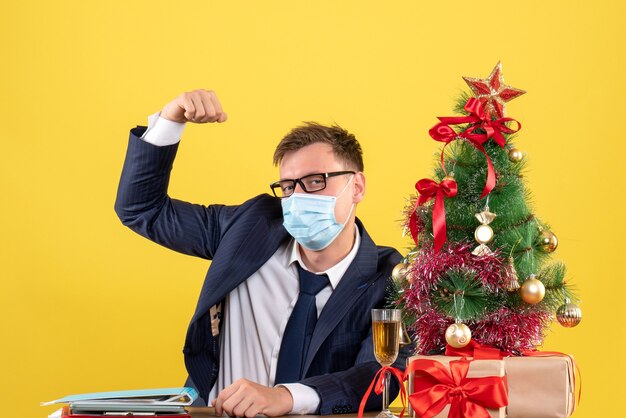 Vista frontal del hombre de negocios que muestra el músculo sentado en la mesa cerca del árbol de Navidad y presenta en amarillo