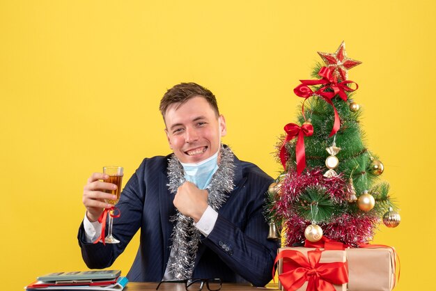 Vista frontal del hombre de negocios quitándose la máscara sentado en la mesa cerca del árbol de Navidad y presenta en amarillo.