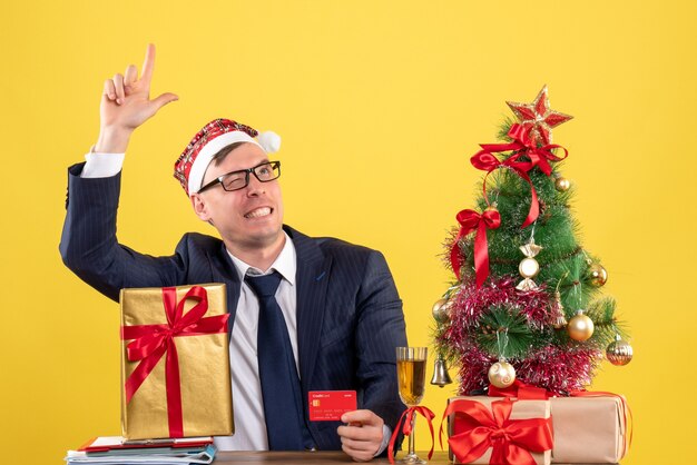 Foto gratuita vista frontal del hombre de negocios con sombrero de santa mirando hacia arriba sentado en la mesa cerca del árbol de navidad y presenta en amarillo