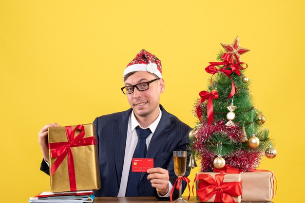 Vista frontal del hombre de negocios con sombrero de santa sentado en la mesa cerca del árbol de Navidad y presenta en amarillo