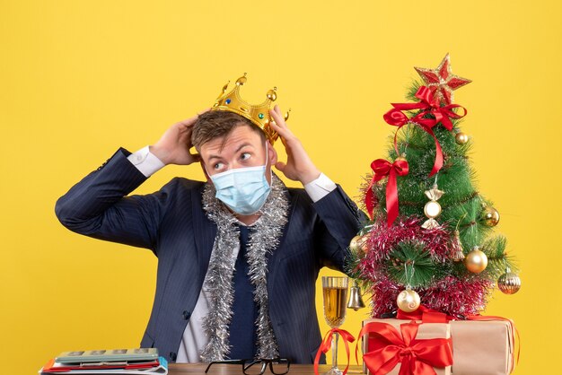 Vista frontal del hombre de negocios sosteniendo su corona sentado en la mesa cerca del árbol de Navidad y presenta en amarillo