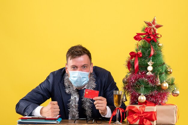 Vista frontal del hombre de negocios con tarjeta de crédito sentado en la mesa cerca del árbol de Navidad y presenta en amarillo