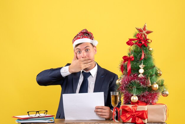 Vista frontal del hombre de ojos abiertos poniendo la mano en la boca sentado en la mesa cerca del árbol de Navidad y presenta en amarillo