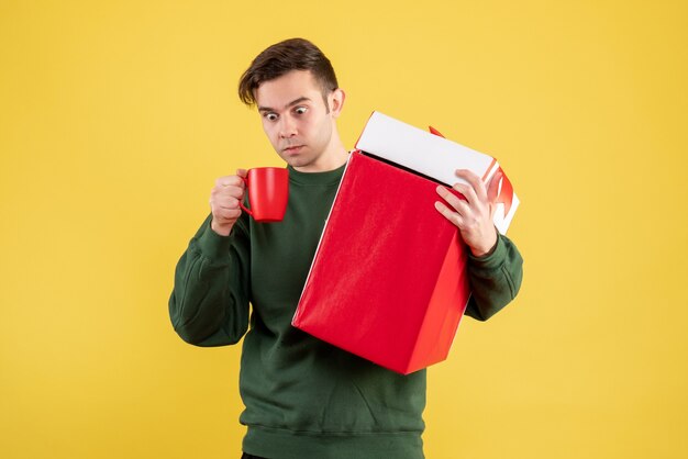 Vista frontal del hombre de ojos abiertos con suéter verde con gran regalo y taza roja de pie sobre amarillo