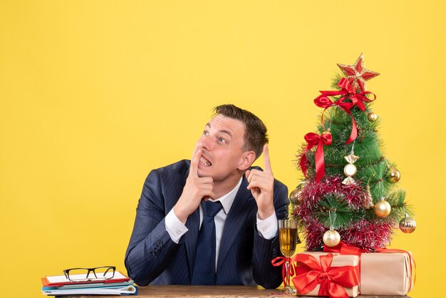 Vista frontal del hombre pensante sentado en la mesa cerca del árbol de Navidad y presenta en amarillo