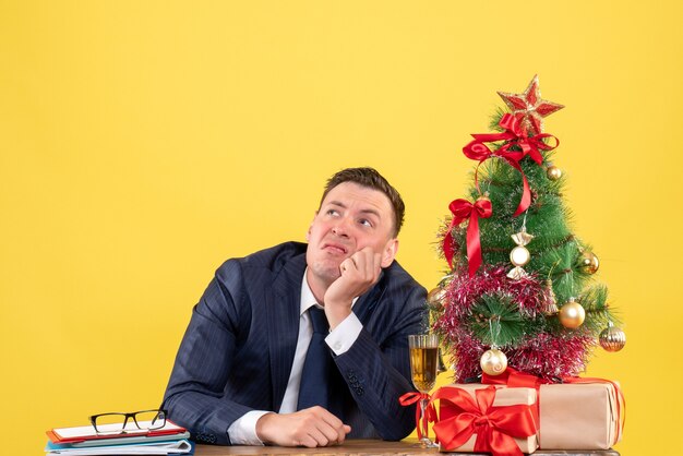 Vista frontal del hombre pensativo mirando alto cartel sentado en la mesa cerca del árbol de Navidad y regalos en amarillo