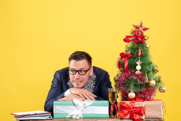 Vista frontal del hombre pensativo poniendo la cabeza en la caja de regalo sentado en la mesa cerca del árbol de Navidad y presenta en amarillo