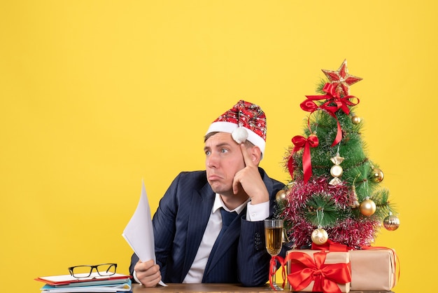 Vista frontal del hombre pensativo sentado en la mesa cerca del árbol de Navidad y presenta en amarillo