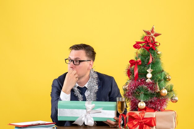 Vista frontal del hombre pensativo sentado en la mesa cerca del árbol de Navidad y presenta en amarillo