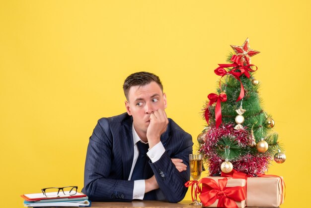 Vista frontal del hombre pensativo sentado en la mesa cerca del árbol de Navidad y presenta en amarillo