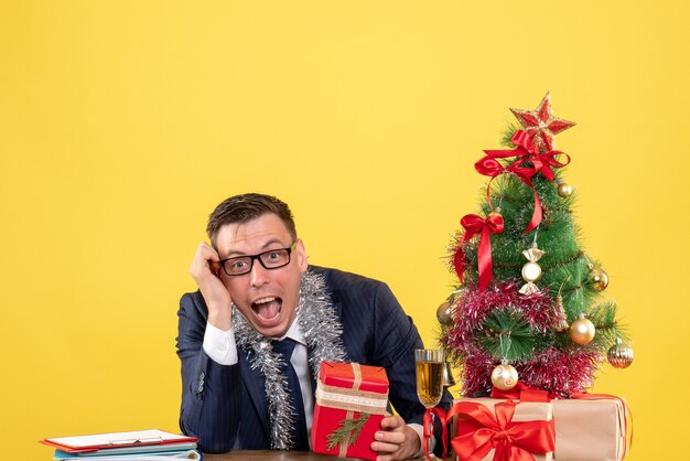 Vista frontal del hombre regocijado con regalo sentado en la mesa cerca del árbol de Navidad y regalos en amarillo