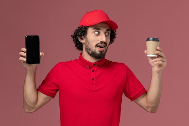 Vista frontal hombre repartidor de mensajería en camisa roja y capa con taza de café de entrega marrón y teléfono en la pared rosa claro empleado de servicio de entrega masculino