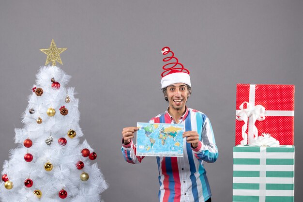 Vista frontal hombre riendo con gorro de Papá Noel de resorte en espiral con mapa cerca del árbol de Navidad blanco y regalos con espacio de copia