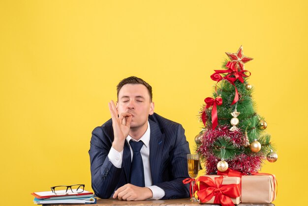 Vista frontal del hombre satisfecho haciendo gesto de beso de chef sentado en la mesa cerca del árbol de Navidad y regalos en amarillo