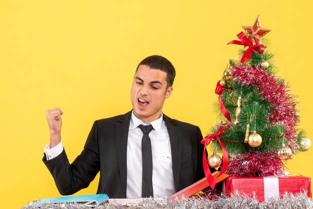 Vista frontal hombre satisfecho sentado en la mesa mostrando gesto ganador árbol de Navidad y regalos