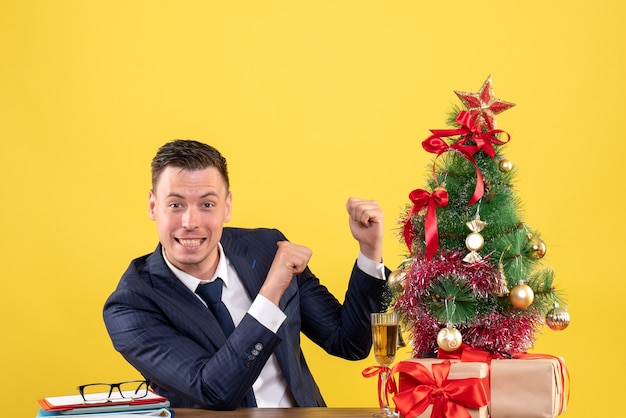 Vista frontal del hombre sonriente apuntando con el dedo hacia atrás sentado en la mesa cerca del árbol de Navidad y regalos en amarillo
