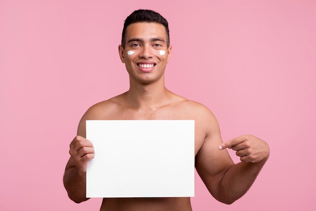 Vista frontal del hombre sonriente con crema en la cara apuntando al cartel en blanco