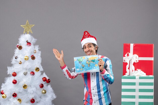 Vista frontal hombre sonriente con gorro de Papá Noel de resorte espiral sosteniendo mapa del mundo apuntando al árbol de Navidad