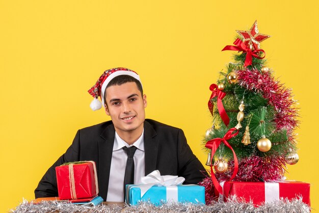 Vista frontal hombre sonriente con gorro de Papá Noel sentado en la mesa con regalos árbol de Navidad y regalos