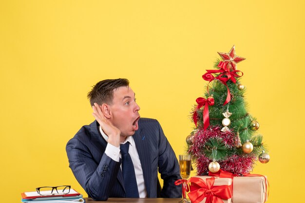 Vista frontal del hombre sorprendido escuchando algo sentado en la mesa cerca del árbol de Navidad y presenta sobre fondo amarillo