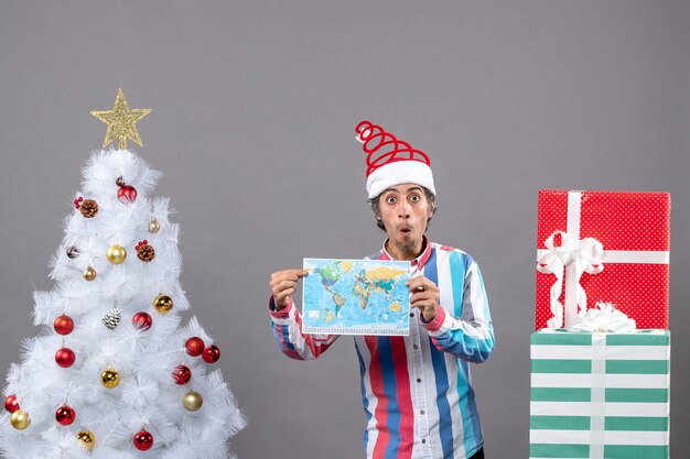 Vista frontal hombre sorprendido con gorro de Papá Noel de resorte en espiral y camisa a rayas con mapa cerca del árbol de Navidad blanco y regalos con espacio de copia