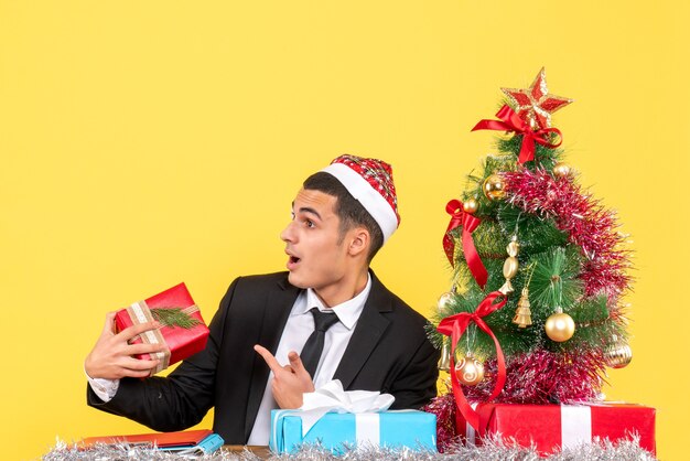 Vista frontal hombre sorprendido con gorro de Papá Noel sentado en la mesa sosteniendo el presente árbol de Navidad y regalos