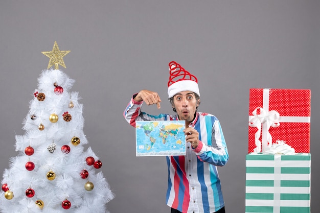 Foto gratuita vista frontal hombre sorprendido con gorro de santa de resorte en espiral y camisa a rayas que muestra el mapa cerca del árbol de navidad blanco y presenta con espacio de copia